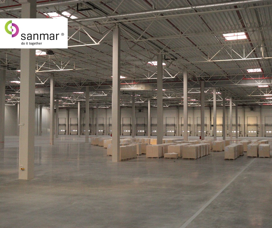 Przestronne wnętrze magazynu z betonową posadzką, rzędem kolumn i bramami w tle. Częściowo załadowane palety z kartonami. Widoczna konstrukcja dachu i oświetlenie.