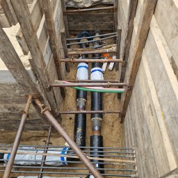 Usługi spawalnicze - Wykonane zabezpieczenie wykopu deskami i metalowymi rozpórkami. Wewnątrz rury, niektóre owinięte folią. Widok z góry na dół, w wykopie z piaskową ziemią.