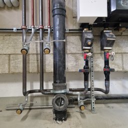 Usługi spawalnicze - Instalacja grzewcza z rurami miedzianymi i czarnymi, zaworami oraz kotłem grzewczym na tle ściany z bloczków. Widoczne elementy mocujące i przyłącza.