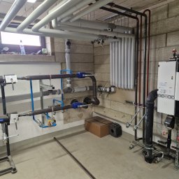 Usługi spawalnicze - Instalacja grzewcza z rurami, zaworami i manometrami w kotłowni. Widoczne elementy spawane i izolowane. Tło stanowi ściana z bloków betonowych.