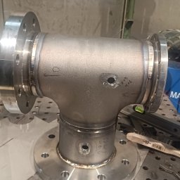 Usługi spawalnicze - Spawany trójnik z kołnierzami, widoczne spawy i otwory. Na stole roboczym leżą narzędzia. Metalowy element po obróbce, gotowy do dalszego montażu.