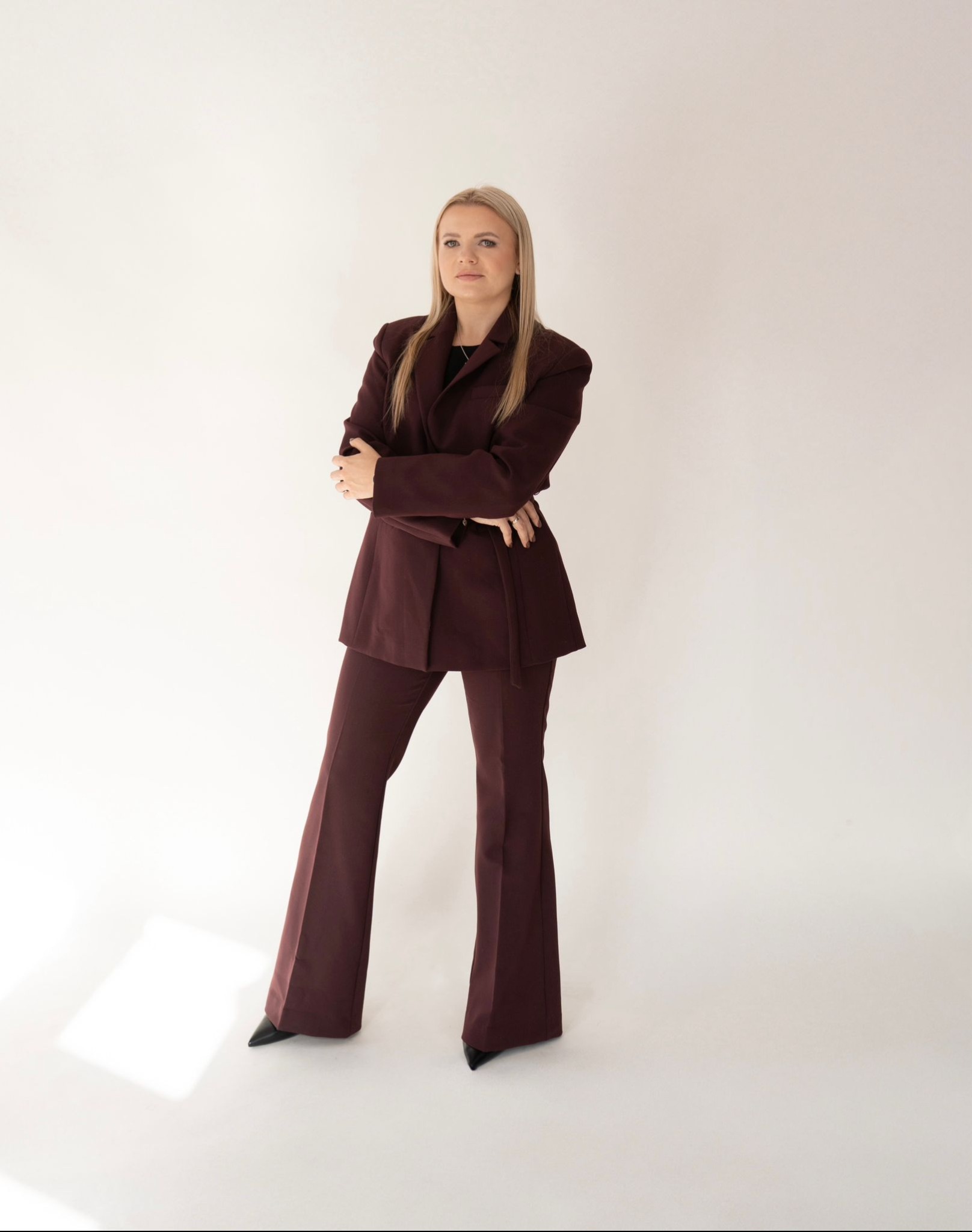 Portret kobiety w bordowym garniturze na jasnym tle. Skrzyżowane ręce, pewny wyraz twarzy. Profesjonalny wizerunek prawnika, radcy prawnego. Cała sylwetka, stonowana kolorystyka.