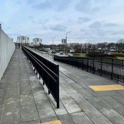 BAZA PROJECT Mariia Sirenko - Zewnętrzna kładka dla pieszych z czarną metalową balustradą i poręczą, wyłożona szarą kostką brukową z żółtymi elementami dla osób niedowidzących. W tle zabudowa miejska.