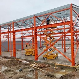 BAZA PROJECT Mariia Sirenko - Konstrukcja stalowa hali w trakcie montażu, pomalowana na pomarańczowo, z widocznymi podnośnikami i pracownikiem na górze. Fundamenty wylane, teren wokół błotnisty.
