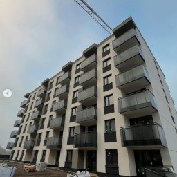 BAZA PROJECT Mariia Sirenko - Nowoczesny budynek wielorodzinny z balkonami o szklanych balustradach, widziany z dołu, z żurawiem budowlanym w tle i materiałami budowlanymi na pierwszym planie.