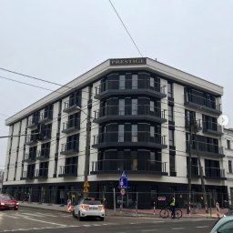 BAZA PROJECT Mariia Sirenko - Nowoczesny budynek apartamentowy 'Prestige' z balkonami z czarnymi balustradami, widok z ulicy, znak drogowy i rowerzysta w tle.