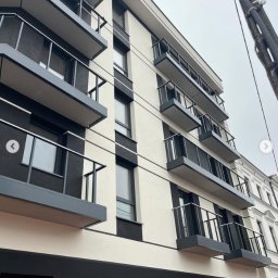 BAZA PROJECT Mariia Sirenko - Nowoczesny budynek z balkonami i balustradami ze stali nierdzewnej, widok z dołu, pochmurne niebo w tle. Minimalistyczny design.