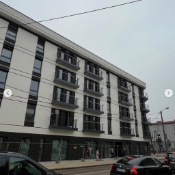 BAZA PROJECT Mariia Sirenko - Nowoczesny budynek wielorodzinny z balkonami zabezpieczonymi metalowymi balustradami. Widok z ulicy, w tle przejeżdżające samochody i pracownik na jednym z balkonów.