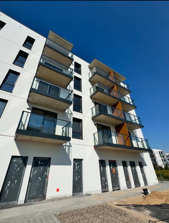 Nowoczesny budynek z balkonami z szklanymi balustradami, widziany z dołu pod kątem, na tle błękitnego nieba. Widoczne zabezpieczone drzwi na parterze.