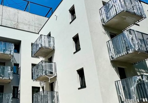 Nowoczesny budynek z balkonami zabezpieczonymi folią ochronną. Czarne ramy okienne kontrastują z białą elewacją. Ujęcie z dołu, widoczne fragmenty niebieskiego nieba.