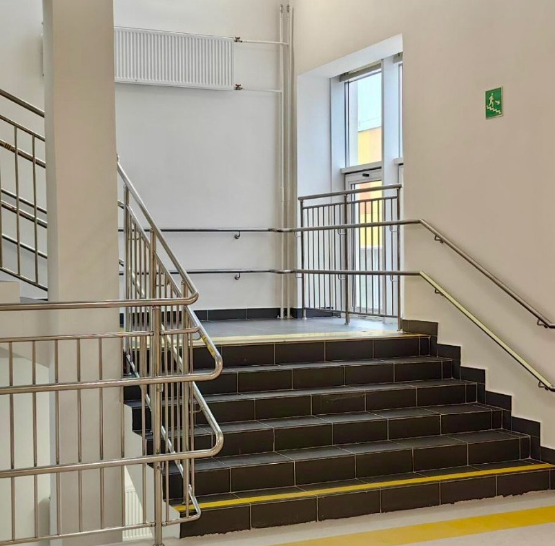 Schody z czarnymi płytkami i metalową balustradą w budynku. Widoczne okno i znak ewakuacyjny. Minimalistyczny design, jasne ściany, dobre oświetlenie.