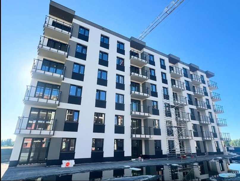 Nowoczesny budynek wielorodzinny z balkonami zabezpieczonymi czarnymi, metalowymi balustradami. Widoczne rusztowania i żuraw budowlany na tle błękitnego nieba.