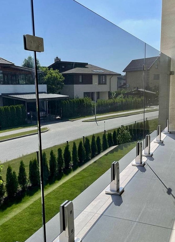 Szklana balustrada balkonowa z metalowymi mocowaniami, widok na zielony trawnik i zabudowę jednorodzinną w słoneczny dzień. Minimalistyczny design.