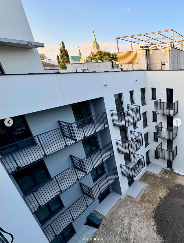 Nowoczesny budynek z balkonami o czarnych, metalowych barierkach, widziany z góry pod kątem. W tle zieleń i wieża kościoła w promieniach zachodzącego słońca.