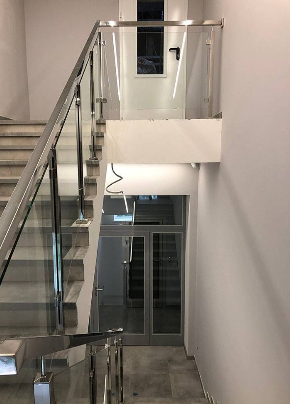 Szklana balustrada ze stalowymi elementami na schodach wewnątrz budynku. Widok z góry na dół, szare płytki na podłodze, białe ściany i drzwi.