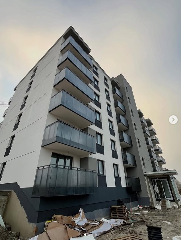 Nowoczesny budynek wielorodzinny z balkonami zabezpieczonymi szklanymi balustradami, widziany z dołu pod kątem. Na pierwszym planie teren budowy.