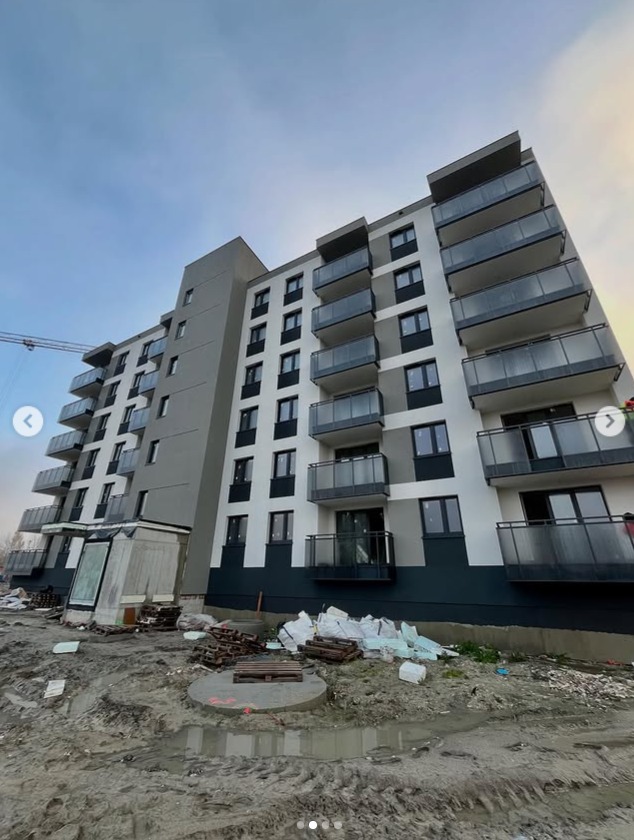 Nowoczesny budynek wielorodzinny z szarymi i białymi elewacjami, balkonami z przeszklonymi balustradami. Ujęcie z dołu, widoczne elementy placu budowy.