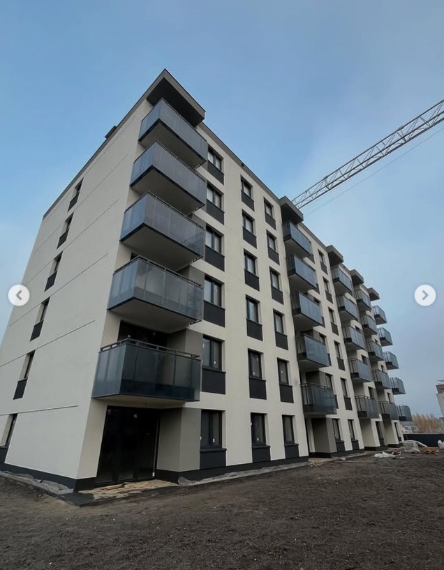 Nowoczesny budynek wielorodzinny z balkonami o szklanych balustradach, widziany z dołu pod kątem. Na górze kadru widoczny fragment żurawia budowlanego.