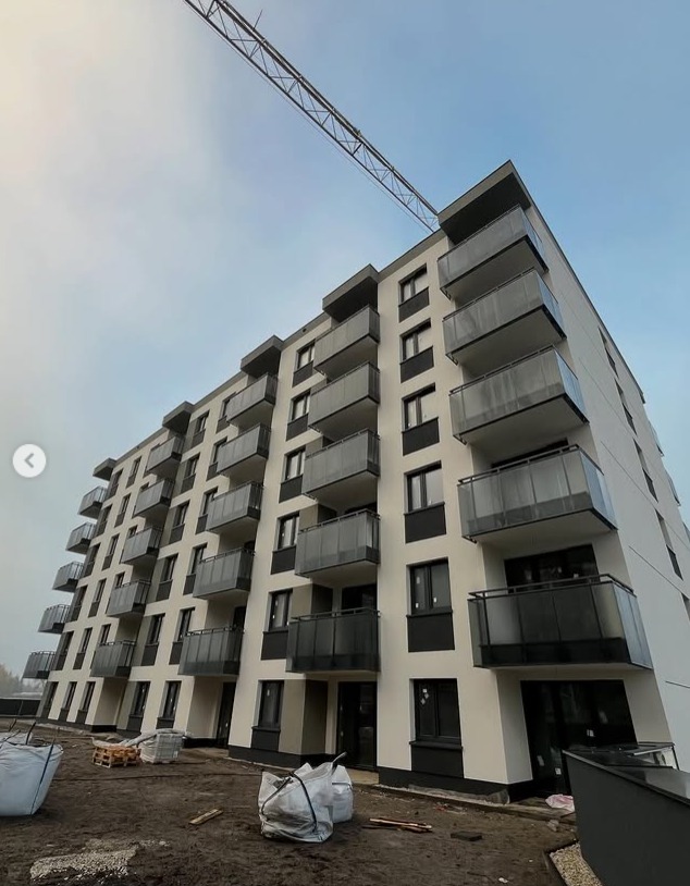 Nowoczesny budynek wielorodzinny z balkonami o szklanych balustradach, widziany z dołu, z żurawiem budowlanym w tle i materiałami budowlanymi na pierwszym planie.