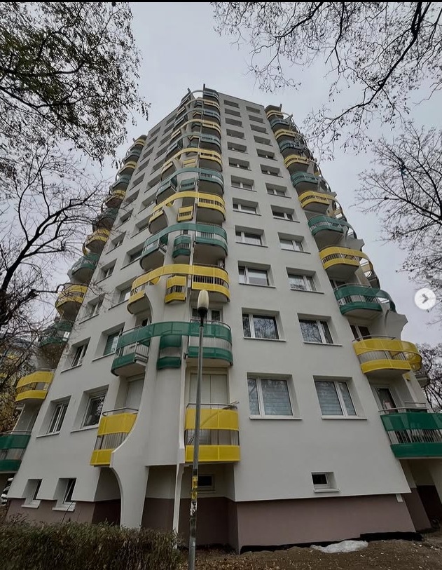 Wysoki blok mieszkalny z kolorowymi balkonami (żółte i zielone) widziany z dołu, z drzewami w tle. Nietypowy kształt balkonów.