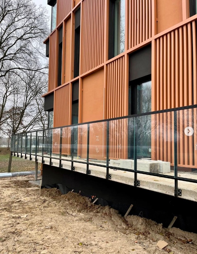 Nowoczesna balustrada szklana z czarnymi elementami mocującymi, zamontowana przy budynku o pomarańczowej elewacji z drewnianymi panelami. Widok z zewnątrz.