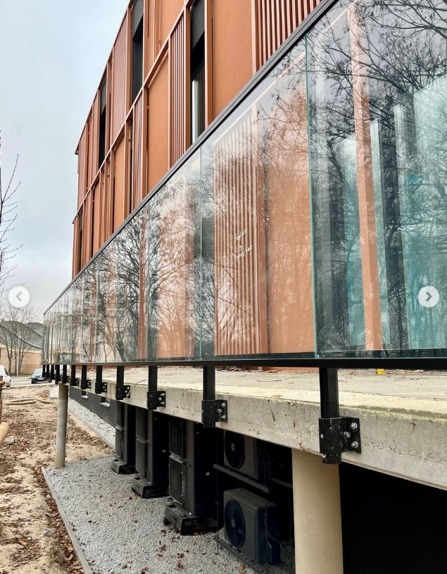 Długa, przeszklona balustrada z czarnymi elementami mocującymi w Nowej Soli. W szkle odbija się las, a w tle widać budynek z brązową elewacją.