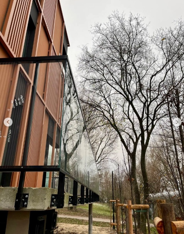 Szklana balustrada z czarnymi elementami mocującymi, zamontowana na betonowej podstawie, z widocznym budynkiem z drewnianą elewacją i drzewami w tle.