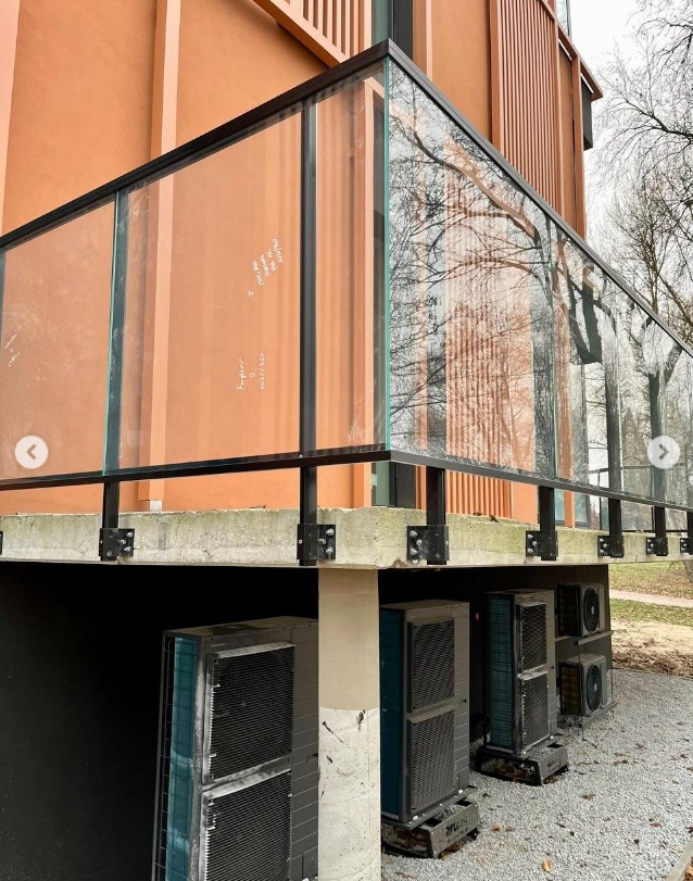 Szklana balustrada z czarnymi elementami mocującymi, zamontowana na betonowej podstawie. W tle budynek z pomarańczową elewacją i widoczne drzewa.