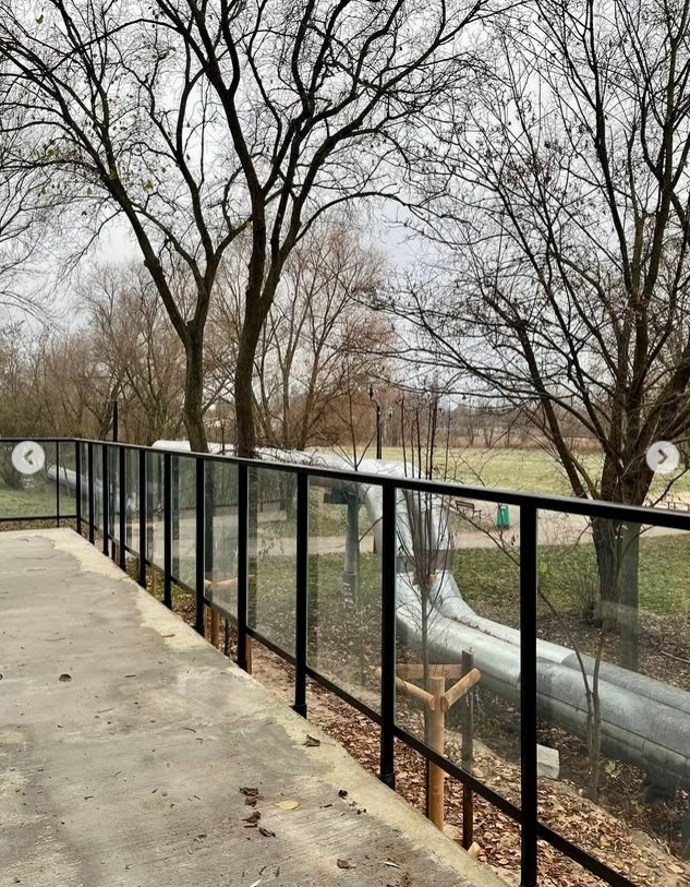 Szklana balustrada z czarnymi, metalowymi słupkami, zamontowana wzdłuż betonowego chodnika w parku. W tle widoczne drzewa i rury ciepłownicze.