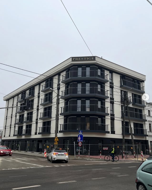 Nowoczesny budynek apartamentowy 'Prestige' z balkonami z czarnymi balustradami, widok z ulicy, znak drogowy i rowerzysta w tle.