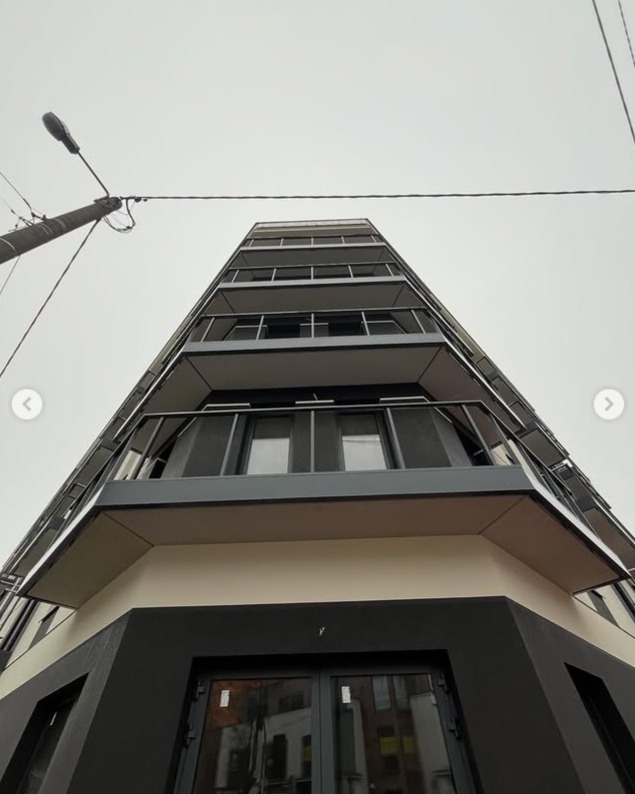 Wysoki budynek z balkonami z metalowymi balustradami, widziany z dołu pod kątem. Minimalistyczna architektura, szara kolorystyka, przewody elektryczne na pierwszym planie.