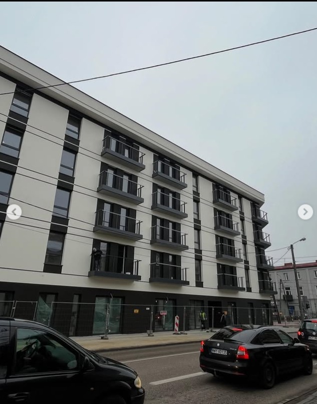 Nowoczesny budynek wielorodzinny z balkonami zabezpieczonymi metalowymi balustradami. Widok z ulicy, w tle przejeżdżające samochody i pracownik na jednym z balkonów.