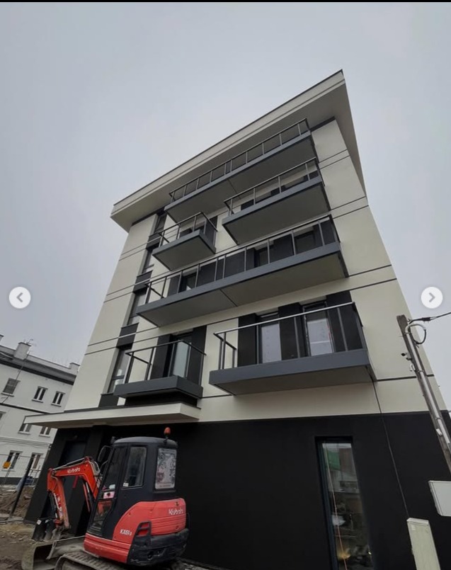 Nowoczesny budynek wielorodzinny z balkonami zabezpieczonymi szklanymi balustradami, widziany z dołu, z małą koparką na pierwszym planie.