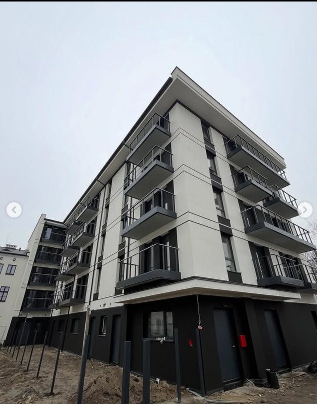 Nowoczesny budynek wielorodzinny z balkonami o stalowych balustradach, widok z dołu na elewację w tonacji bieli i szarości, pochmurne niebo w tle.