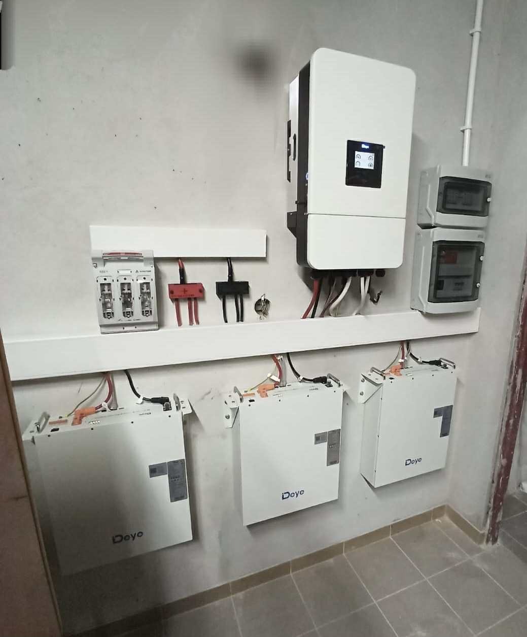 Instalacja magazynu energii Deye: trzy białe moduły na ścianie, inwerter, zabezpieczenia i skrzynka elektryczna. Widok na szarą ścianę i podłogę z płytek.
