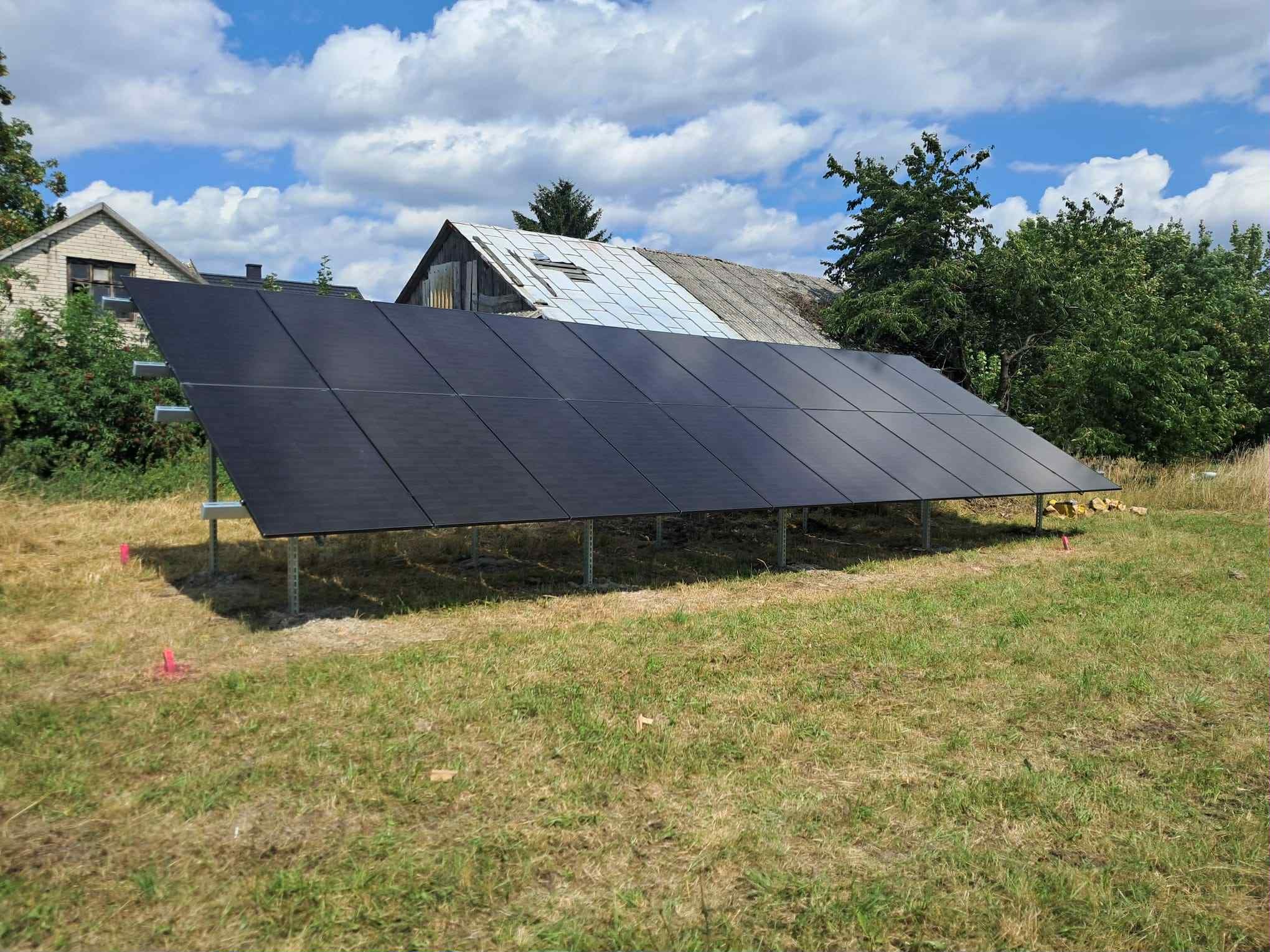 Czarny panel fotowoltaiczny na metalowej konstrukcji, ustawiony na trawie przed budynkiem gospodarczym z częściowo odnowionym dachem w słoneczny dzień.