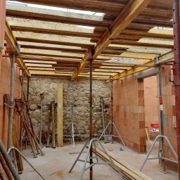 Wnętrze budynku w trakcie budowy: murowane ściany z pustaków, drewniane szalunki stropu podparte metalowymi stemplami, fragment ściany z kamienia.