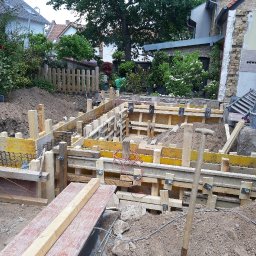 CIRO BAU SPÓŁKA Z OGRANICZONĄ ODPOWIEDZIALNOŚCIĄ - Szczegółowy widok szalunku fundamentów zbrojonych stalą, przygotowanego do zalania betonem, na tle zieleni i fragmentu budynku w trakcie remontu.