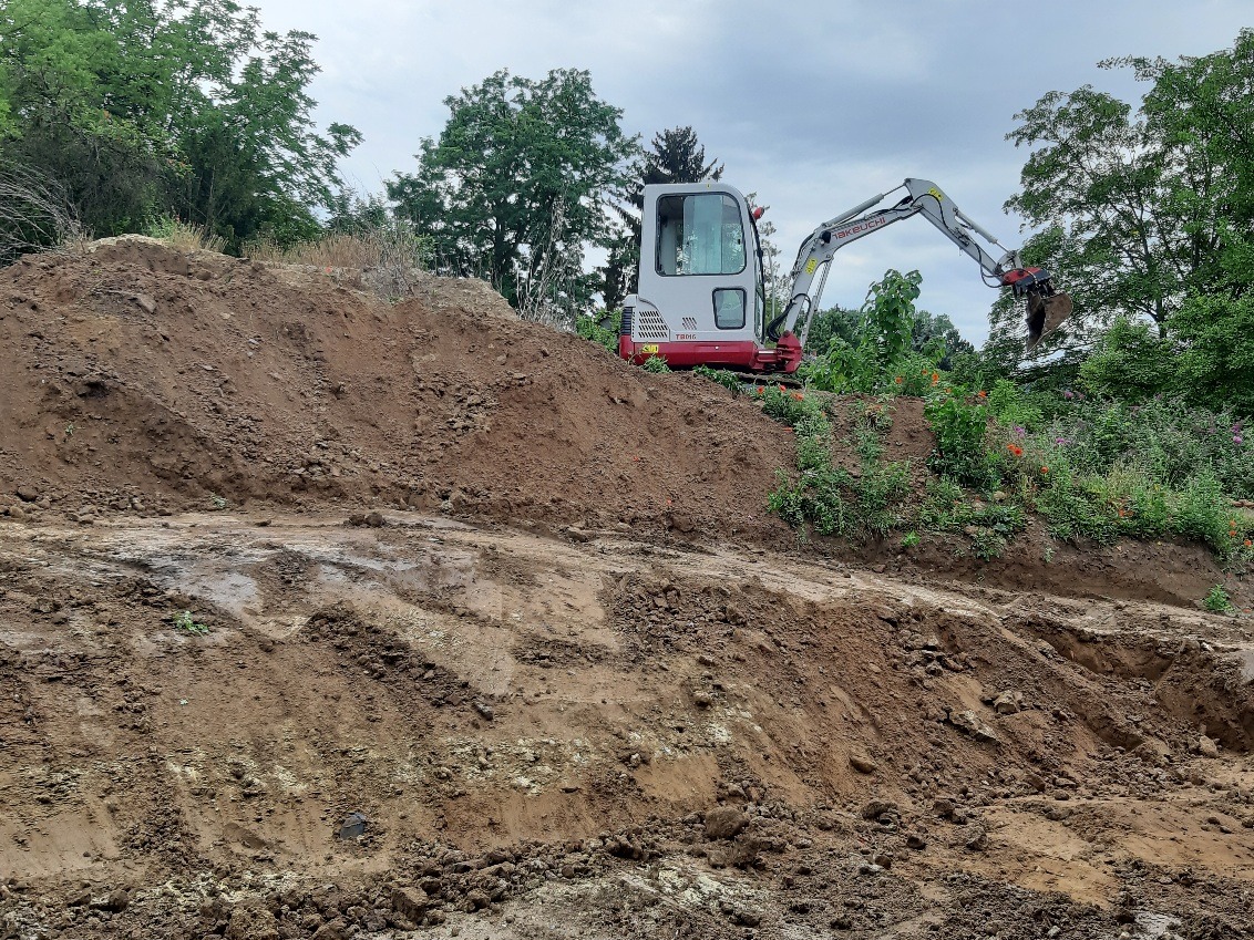 Mini koparka Takeuchi TB016 na nierównym terenie z roślinnością, w trakcie prac ziemnych. Widoczne ślady opon i świeżo przekopana ziemia.