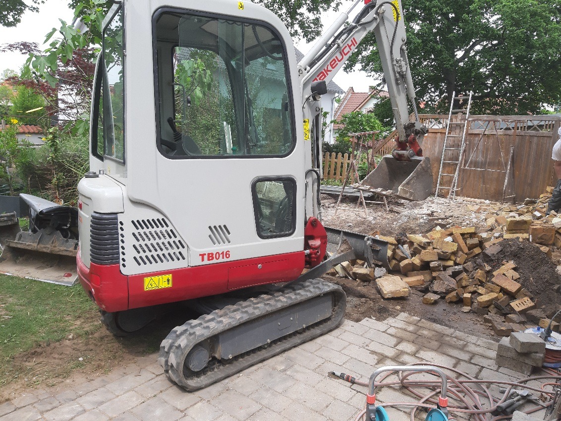 Mini koparka Takeuchi TB016 na posesji z gruzem i ziemią, widoczne ogrodzenie i drabina. Prace ziemne w ogrodzie, sprzęt budowlany w akcji.