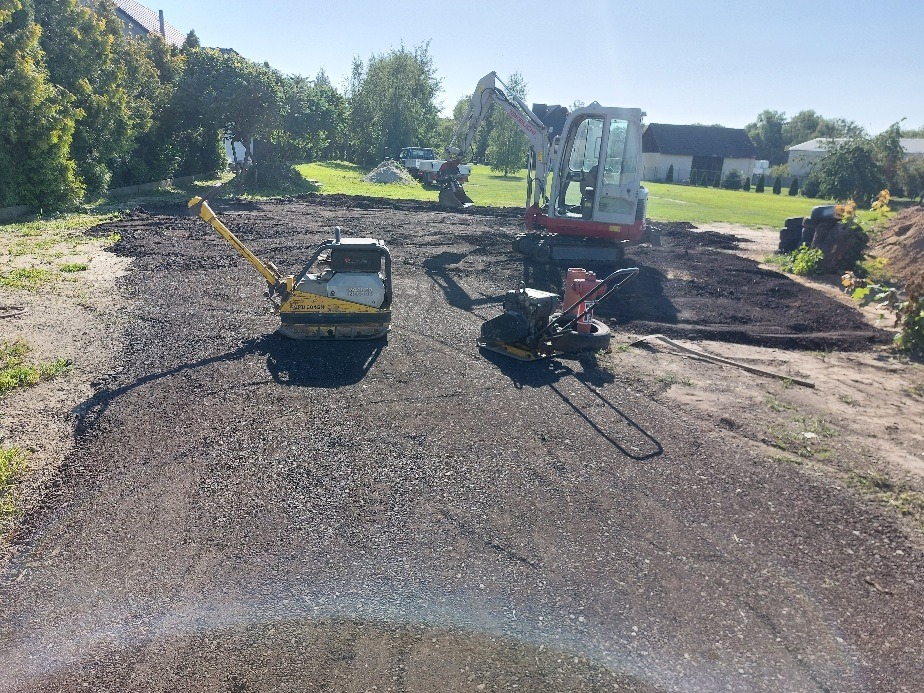 Utwardzanie terenu: zagęszczarka, walec i koparka na placu budowy. Widoczne świeżo rozłożone warstwy kruszywa. Prace ziemne w toku, słoneczny dzień.