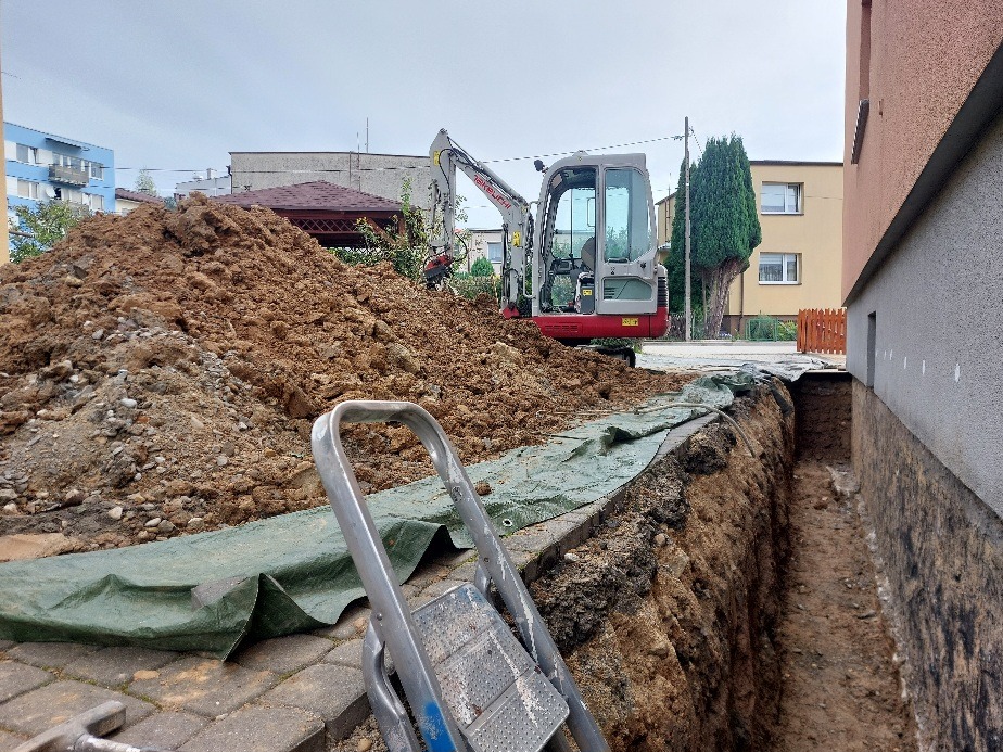 Wąski wykop wzdłuż ściany budynku, zabezpieczony zieloną folią. W tle minikoparka i sterta ziemi. Na pierwszym planie fragment drabiny.