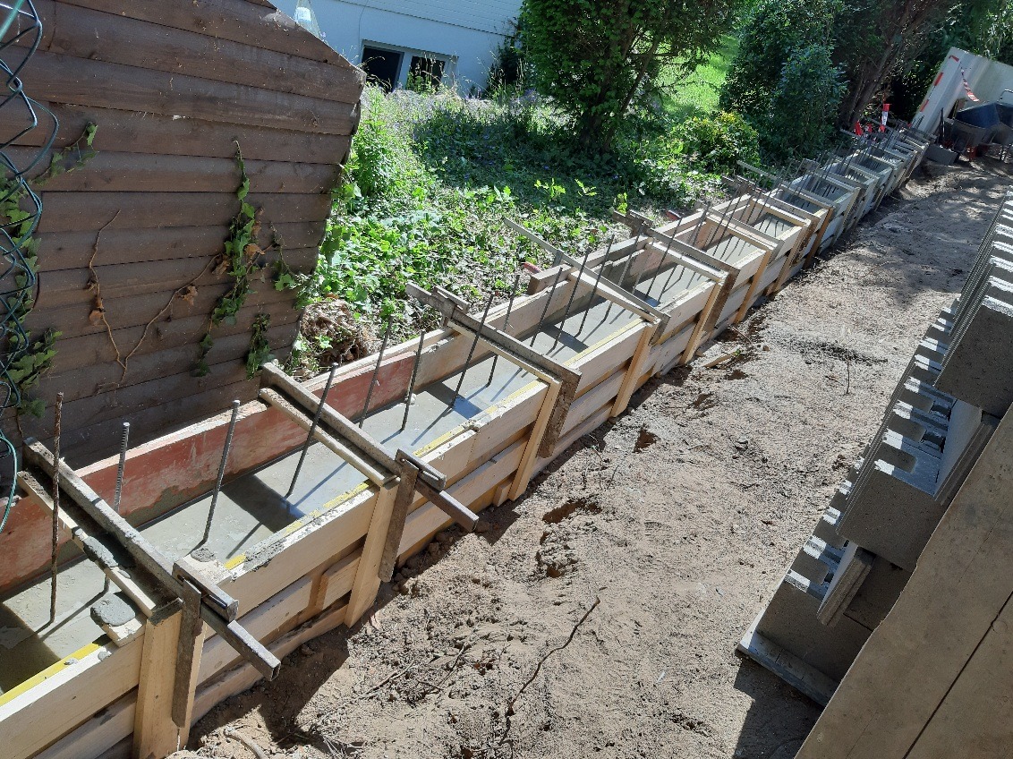Zalewanie betonem szalunku zbrojonego prętami, przygotowanego pod fundament. Widoczne deski szalunkowe i świeży beton. Częściowo ułożone bloczki fundamentowe.