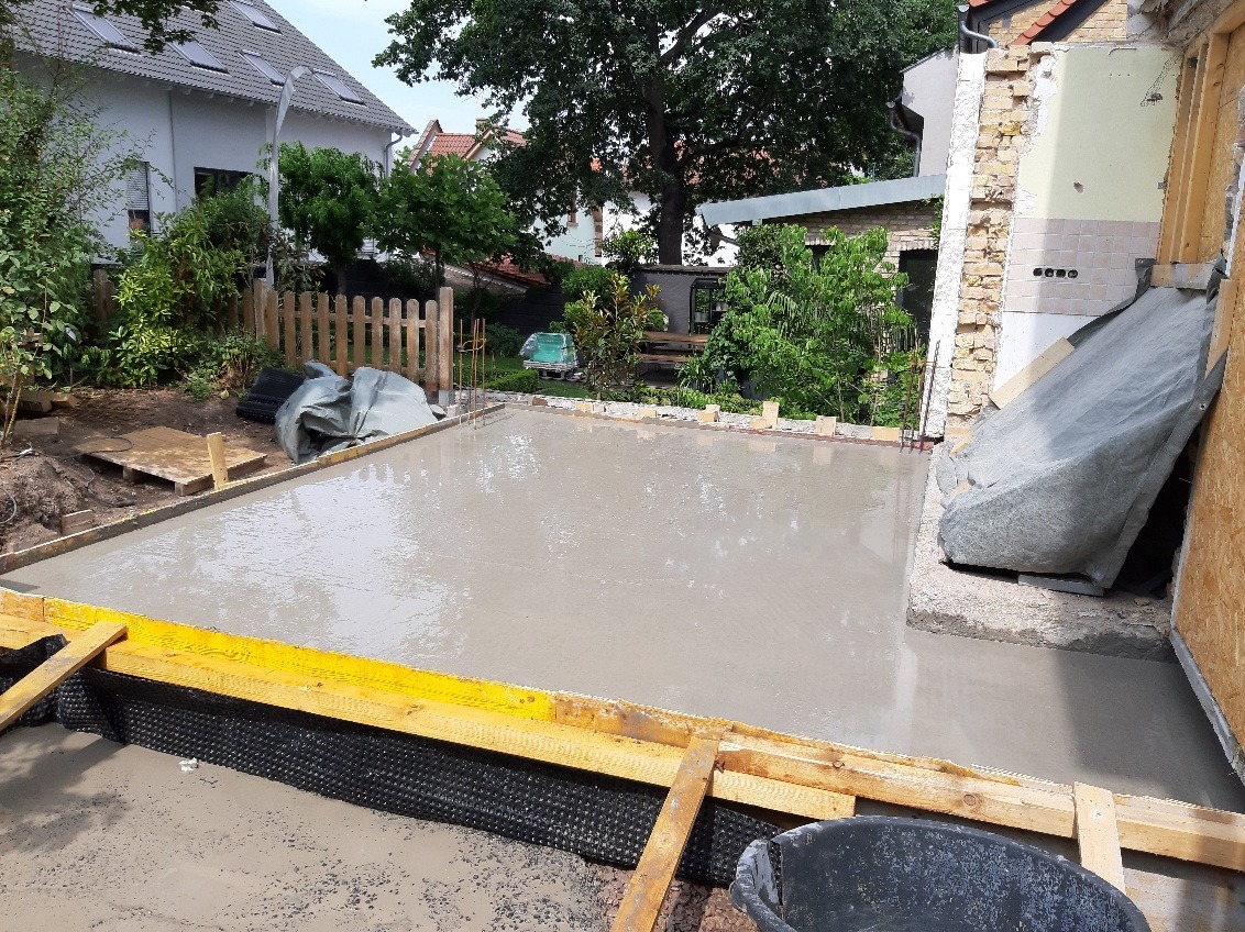 Wylany beton na przygotowanym fundamencie z żółtym szalunkiem i czarną folią izolacyjną. W tle zieleń ogrodu i zabudowania.