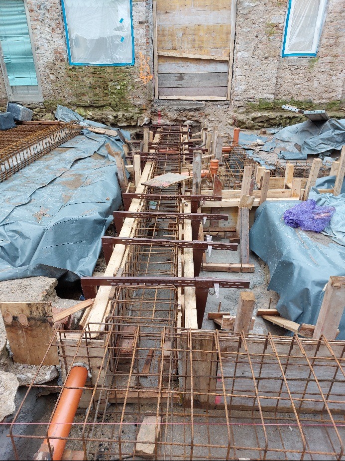 Zbrojenie fundamentów w wykopie, widoczne drewniane szalunki i rury kanalizacyjne. W tle fragment starego budynku z zamurowanymi oknami.