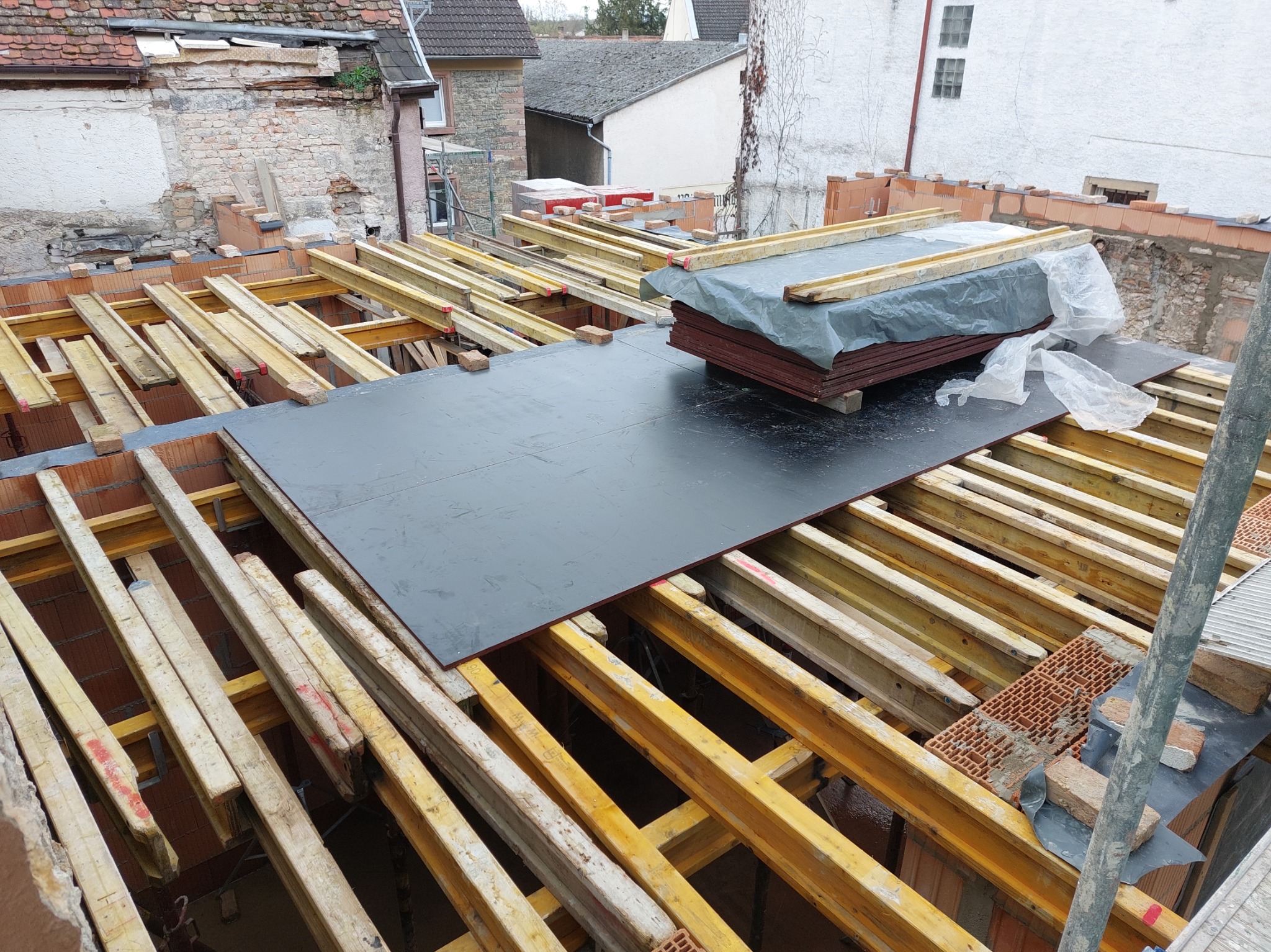 Konstrukcja stropu z drewnianych belek i płyt szalunkowych na budowie domu, widok z góry. Częściowo wymurowane ściany z czerwonej cegły. W tle zabudowa mieszkalna.