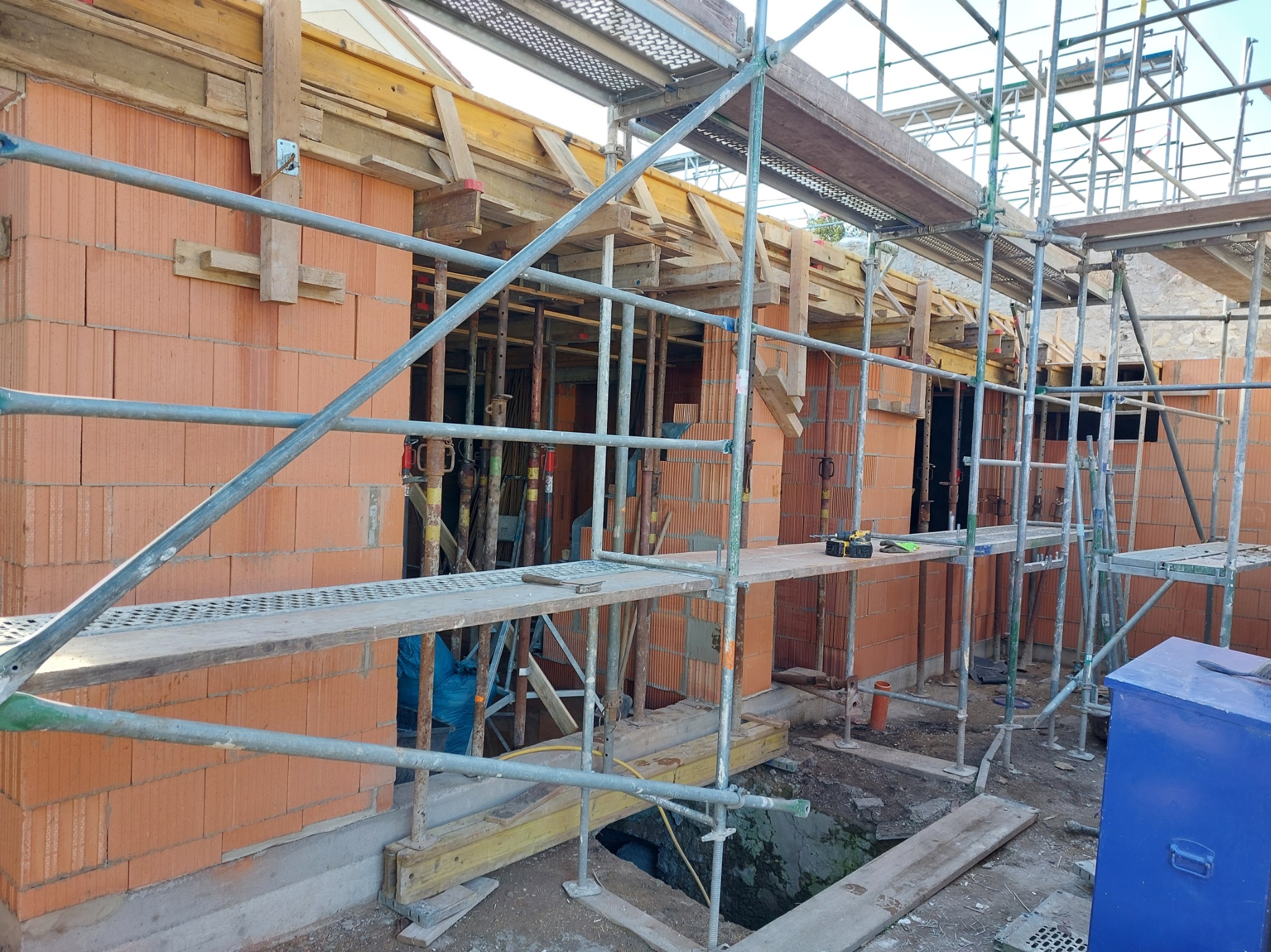 Wznoszenie ścian z czerwonej cegły na budowie. Widoczne rusztowania, drewniane elementy konstrukcyjne i wykop. Cegły z charakterystycznymi otworami.