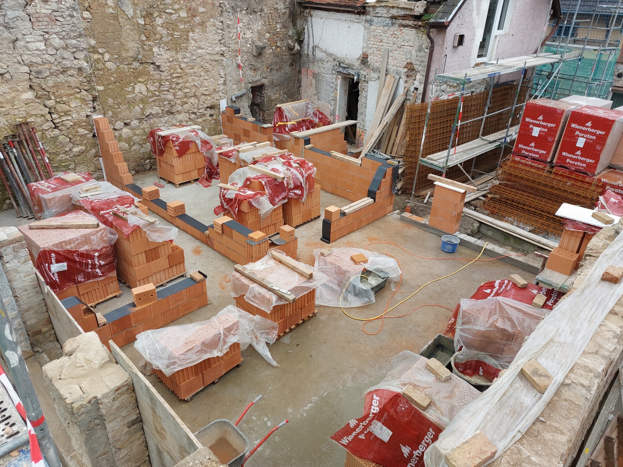 Wysoki kąt na plac budowy z częściowo wzniesionymi ścianami z czerwonej cegły Poroton, zabezpieczonymi folią, z widocznymi rusztowaniami i starym murem w tle.