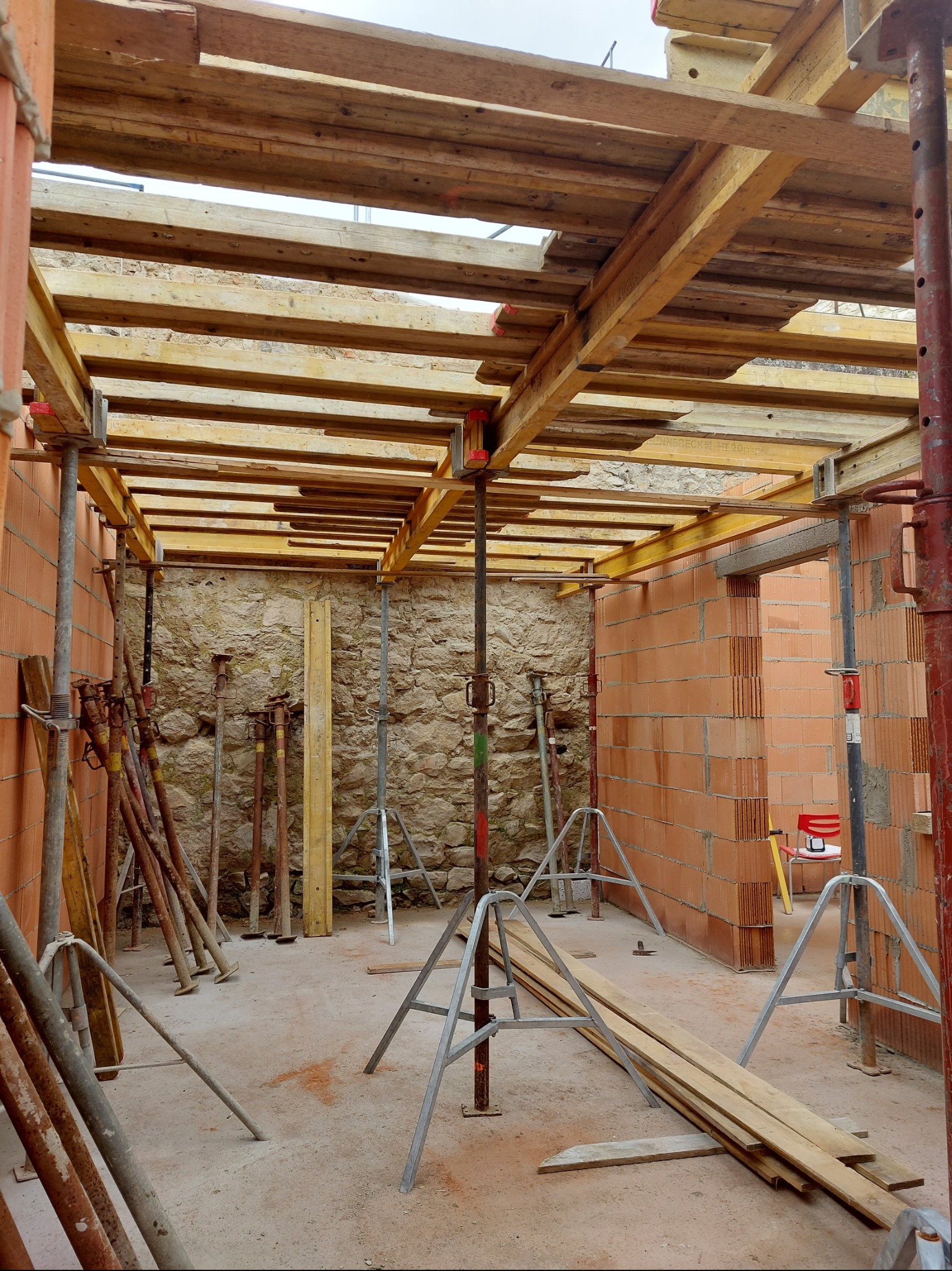 Wnętrze budynku w trakcie budowy: murowane ściany z pustaków, drewniane szalunki stropu podparte metalowymi stemplami, fragment ściany z kamienia.