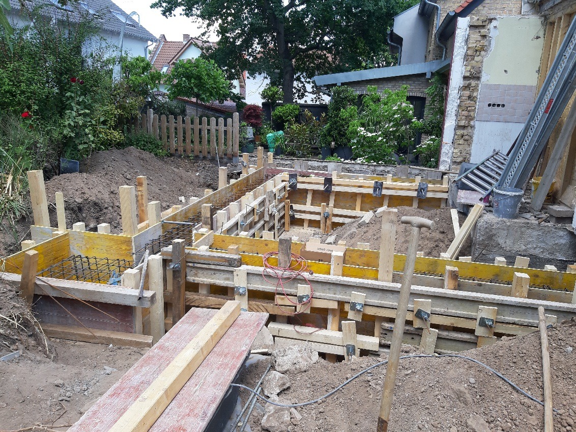 Szczegółowy widok szalunku fundamentów zbrojonych stalą, przygotowanego do zalania betonem, na tle zieleni i fragmentu budynku w trakcie remontu.